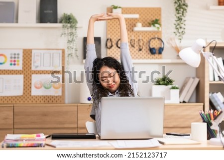 Asian girl finished computer work,  stretching relaxing seated at workplace feeling stress relief, satisfied by project accomplishment, task done in time. Long curly hair woman happy and relaxed.