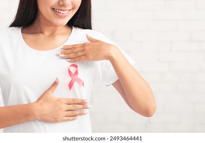 Asian Girl Framing Cancer Ribbon Symbol On Pink T-Shirt Posing Against White Brick Wall Indoors. Cropped - Powered by Shutterstock