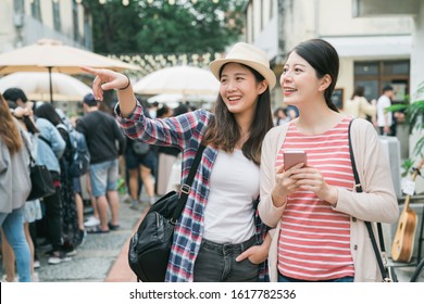 Asian Girl Friends At Farmer Market Outdoors In Summer In City. Two Young Female Travelers Holding Cellphone With Map Online App Searching And Point Finger While Find Shop Destination Tokyo Japan