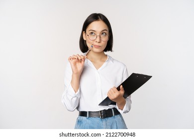 Asian girl in glasses thinks, holds pen and clipboard, writing down, making notes, standing over white background - Shutterstock ID 2122700948