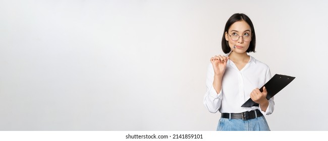 Asian girl in glasses thinks, holds pen and clipboard, writing down, making notes, standing over white background