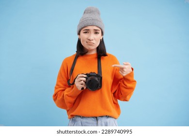 Asian girl in hat, points at her digital camera with disappointed, upset face, doesn't like her device, photographer complains at her digicam, blue background.