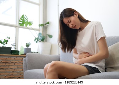 Asian girl having period is sitting on sofa and feeling much of painful on her stomach which called women's pain or menstrual syndrome. She holds onto her stomach and curls through spasms. 