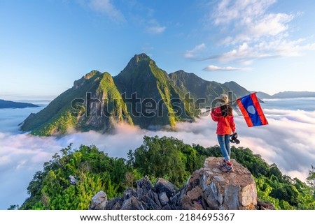 Asian girl holding Laos flag on top of Viewpoint of Nong Khiaw - a secret village in Laos. Stunning scenery of limestone cliff valley covered with fog
