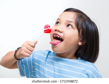 Asian Girl Kid Consuming Sweet Candy Lollipop With Sugar Added, Causing Loss Teeth And Unhealthy Oral Care. Funny Portrait Of Girl Eating Sugary Food On White Background.