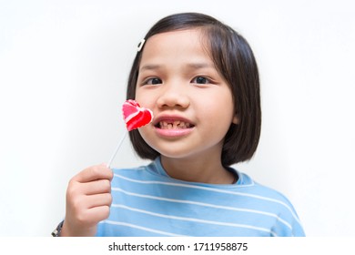 Asian Girl Kid Consuming Sweet Candy Lollipop With Sugar Added, Causing Loss Teeth And Unhealthy Oral Care. Funny Portrait Of Girl Eating Sugary Food On White Background.