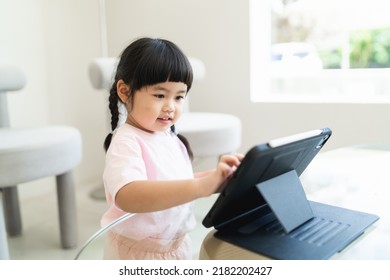 Asian Girl Looking And Touch On Tablet Screen Attentively. Overstimulated Children Concept. Too Much Screen Time. Asian Girl Watching Videos While Tv Is Working.