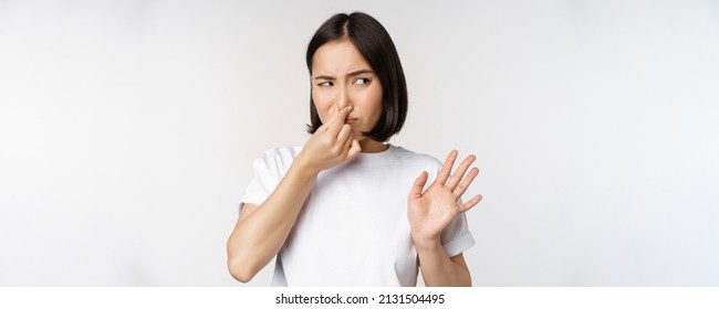 Asian Girl Looks Disgusted, Rejecting Product With Bad Smell, Shut Nose From Aversion And Cringe, Standing Against White Background