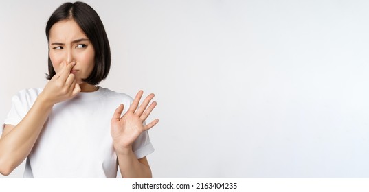 Asian Girl Looks Disgusted, Rejecting Product With Bad Smell, Shut Nose From Aversion And Cringe, Standing Against White Background