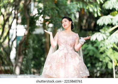 Asian Girl In An Old Rose Pink Color Knee Length Fluffy Dress With High Heel Slingback Shoes And Ceremonial Thread On Her Wrist. She Is Brighter Mood  Posing In The Shade Of Tree Garden.