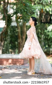 Asian Girl In An Old Rose Pink Color Knee Length Fluffy Dress With High Heel Slingback Shoes And Ceremonial Thread On Her Wrist. She Is Brighter Mood  Posing In The Shade Of Tree Garden.