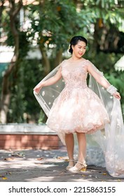 Asian Girl In An Old Rose Pink Color Knee Length Fluffy Dress With High Heel Slingback Shoes And Ceremonial Thread On Her Wrist. She Is Brighter Mood  Posing In The Shade Of Tree Garden.