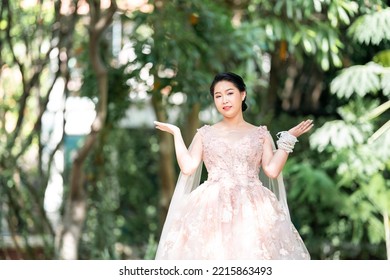 Asian Girl In An Old Rose Pink Color Knee Length Fluffy Dress With High Heel Slingback Shoes And Ceremonial Thread On Her Wrist. She Is Brighter Mood  Posing In The Shade Of Tree Garden.