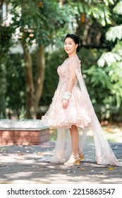 Asian Girl In An Old Rose Pink Color Knee Length Fluffy Dress With High Heel Slingback Shoes And Ceremonial Thread On Her Wrist. She Is Brighter Mood  Posing In The Shade Of Tree Garden.