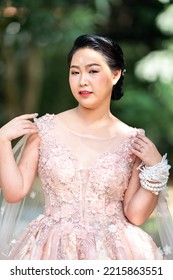 Asian Girl In An Old Rose Pink Color Knee Length Fluffy Dress With High Heel Slingback Shoes And Ceremonial Thread On Her Wrist. She Is Brighter Mood  Posing In The Shade Of Tree Garden.