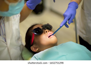 Asian Girl Patient Lying Down In Clinic During Appointment.Nurse's Hand With Purple Glove Sucking Water From Mouth Using Machine During Surgery.Young Child Having Dental Check While Dentist Looks On.