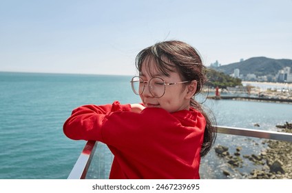 Asian girl in a red sweater relaxes by the sea railing, savoring the coastal scenery - Powered by Shutterstock