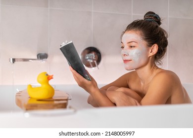 Asian Girl Running A Bath At Home Taking Relaxing Time For Herself In Cozy Winter Apartment Applying Clay Mask And Reading A Book Soaking In Hot Water With Rubber Duck Toy On Bathtub Caddy