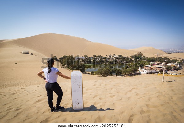 Asian girl with sand board looking down at\
hidden oasis standing in sand dunes\
desert.