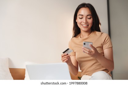 Asian Girl Shops Online From Home. Young Woman Sitting On Bed With Credit Card, Smartphone And Laptop. Female Order On Mobile Phone App, Paying In Internet