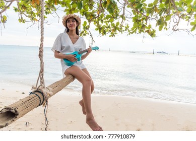 An Asian girl sits on a swing at the beach with an instrumental. Ukulele is a classic song, dressed in a solitary white outfit and reminiscent of a lover far away in summer.