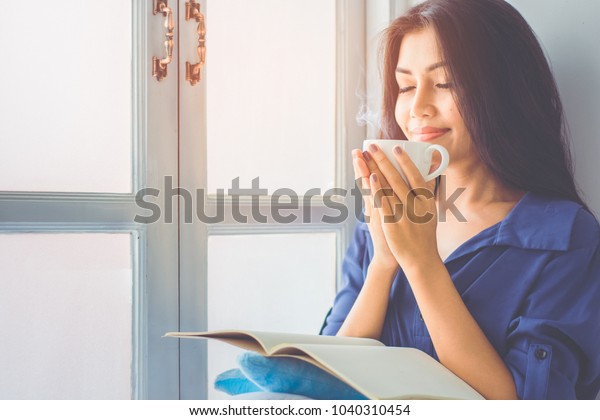 Asian girl sitting by the window. She is smiling\
and holding a cup of coffee in hand split up in order to inhale the\
aroma of coffee. Amidst the mild morning sun shines through the\
glass window.