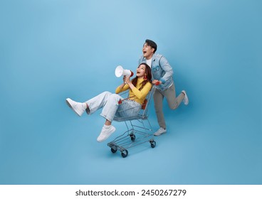 Asian girl sitting inside of shopping trolley and holding megaphone and Asian man pushing shopping cart running to supermarket isolated on blue background. - Powered by Shutterstock