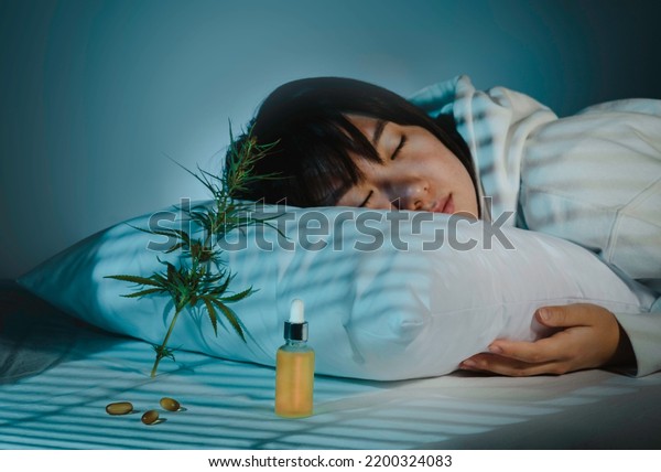 Asian girl sleeping in evening bedroom with cbd\
oil, capsules and a cannabis branch. Melatonin production, concept\
of combat sleep disorders