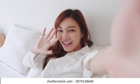 ฺBeautiful Asian girl smiling and show her hand in a white shirt takes a video selfie with a friend on the bed in a white room. - Powered by Shutterstock