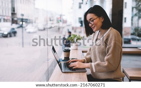 Asian girl typing and watching on laptop computer in cafe. Young concentrated millennial brunette woman wearing glasses sitting at table near window. Freelance and remote work. Modern female lifestyle