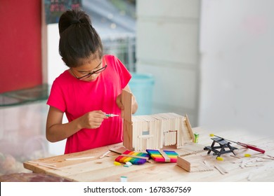 Asian Girl Using Glue To Fix Popsicle Sticks Project. Child Crafting Using Sticks.Young Female Homeschooler Making Craft Using Wooden Stick On Table In Outdoor.Mini Picnic Bench.Homeschooling.