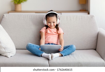 Asian Girl Using Tablet Computer Wearing Wireless Headphones Watching Cartoons Sitting On Couch At Home. Kid And Gadget Concept
