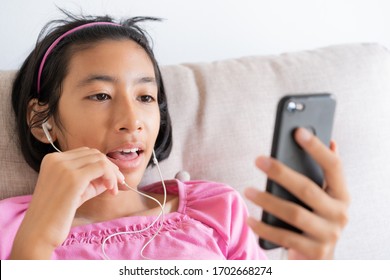 Asian Girl Video Call On The Smartphone While Sitting On The Sofa In The Living Room At Home, Happy Asia Child Smile On The Face And Chat With Earphone Past Internet. Lifestyle And Relaxing Concept