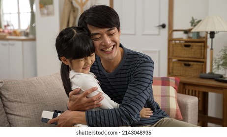 Asian Girl Walking Towards Dad And Showing Him Surprise Gift On Father’s Day At Home. The Touched Man Giving His Daughter A Big Hug On The Sofa