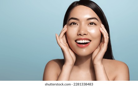 Asian girl washes her face, smiles and applied cosmetic product. Young woman using vitamin a serum, rubbing cream on her facial skin, has glowing body, standing over blue background. - Powered by Shutterstock