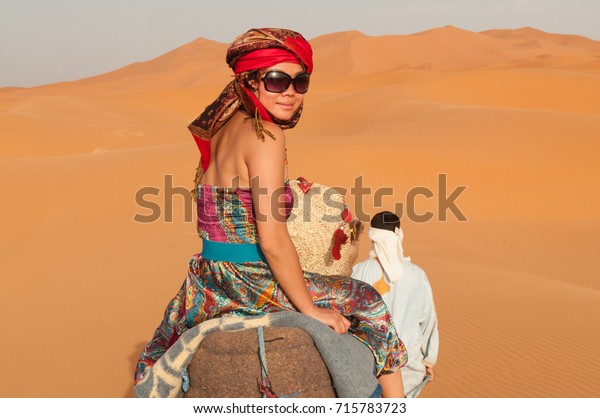 Asian girl wearing colourful\
clothes, a head scarf and harem pants riding a camel across the\
Sahara Desert in Morocco, North Africa, turns back to look at the\
camera.