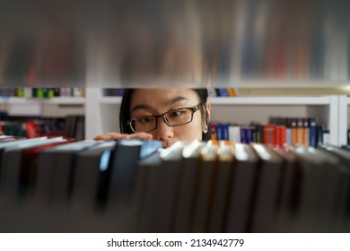 Asian girl wearing glasses standing between bookshelves in university library, picking up book from shelf, female student choosing textbook, looking for literature while preparing for exam. Education - Powered by Shutterstock