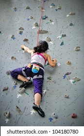 Asian Girl Wearing Harness And Belaying Rope, Climbing On A Very High Rock Climbing Wall.