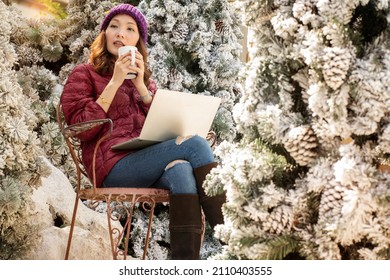 Asian girl wearing a jacket, knitting hat, drinking coffee, breakfast and checking emails, working with laptops in the pine tree garden. It's cold in the snow. Sit and work in the garden. 