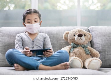 Asian girl wearing mask hygienic stay isolation at home for self quarantine for protect against virus COVID-19,little girl sitting play mobile living on sofa at home with teddy bear friend beside. - Powered by Shutterstock