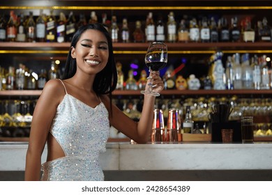 Asian girl wearing a white dress smiles happily, holding a glass of red wine, standing in front of the bartender's drinks counter in a nightclub. - Powered by Shutterstock