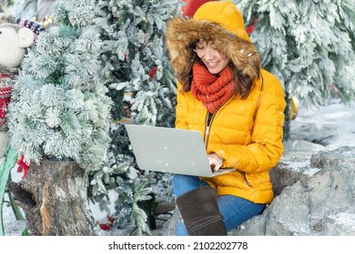 Asian girl wearing a yellow jagger, sitting in a pine garden in winter snow and working online with laptop .Work anywhere concept.