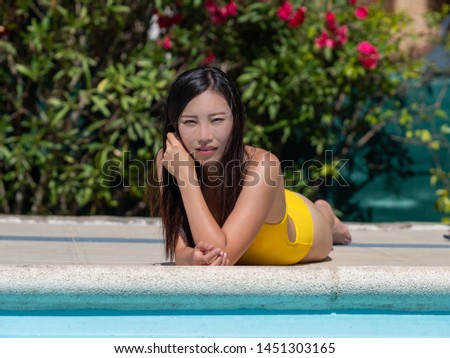 Asian girl in yellow bikini takes the sun and interacts with her mobile phone next to a swimming pool