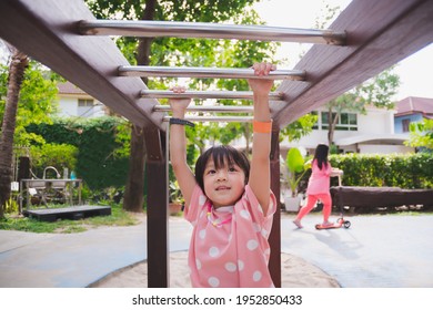 Asian girls are playing on playground with trapeze. Active child hangs on bar with two hands. Children exercise. Behind the child is riding scooter. Sink clean hands are on the side. In summer time.