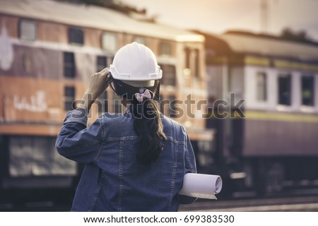 Asian girls train engineer, dressed in jeans and wearing a White Helmet are standing looking at the moving train station amidst the evening sun.