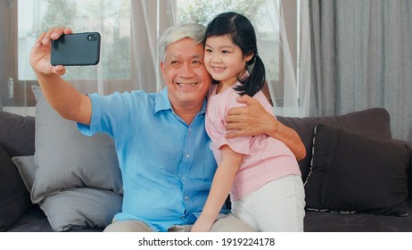 Asian Grandfather And Granddaughter Video Call At Home. Senior Chinese Grandpa Happy With Young Girl Using Mobile Phone Video Call Talking With Her Dad And Mom Lying In Living Room At Home.