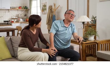 Asian Grandfather Holding His Knee Joint And Groaning In Pain While Standing Up From Couch. His Adult Granddaughter Comes Over To Support Him And Massage His Legs