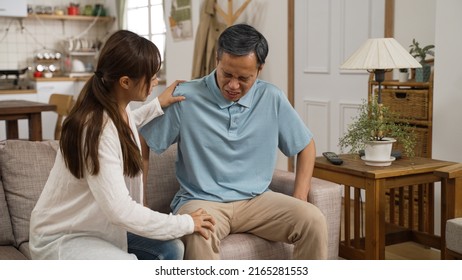 Asian Grandfather Holding His Knee Joint And Groaning In Pain While Standing Up From Couch. His Adult Granddaughter Comes Over To Support Him And Massage His Legs