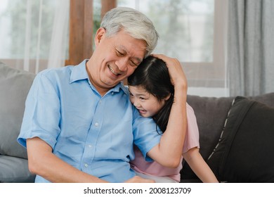 Asian grandfather talking with granddaughter at home. Senior Chinese, grandpa happy relax with young granddaughter girl using family time relax with young girl kid lying on sofa in living room.