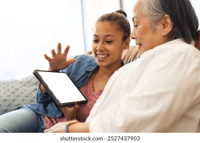 Asian grandmother and biracial teenage granddaughter sitting at home, looking at tablet with copy space. Grandmother has grey hair, granddaughter has light brown hair, unaltered - Powered by Shutterstock